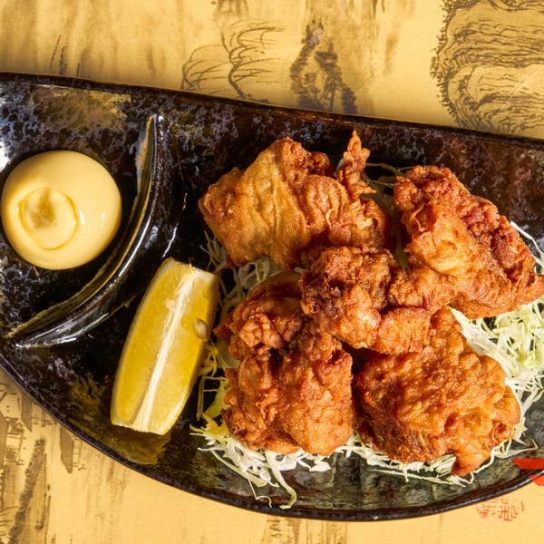 Crispy fried chicken karaage with shredded cabbage, a lemon wedge, and creamy sauce on a black plate.