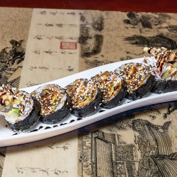 A white platter holds a row of sushi rolls topped with sesame seeds and sauce on a patterned tablecloth.