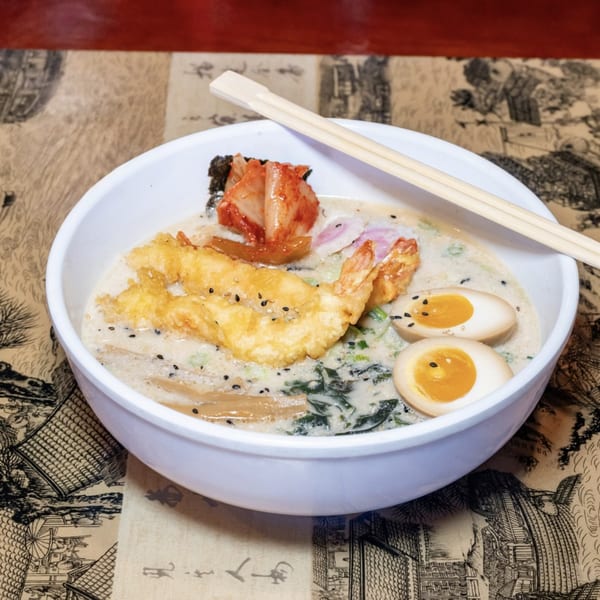 Creamy ramen with tempura shrimp, kimchi, soft-boiled eggs, and chopsticks in a white bowl.