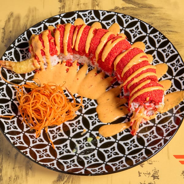 Red sushi roll with orange sauce and fried shrimp on a black and white patterned plate.
