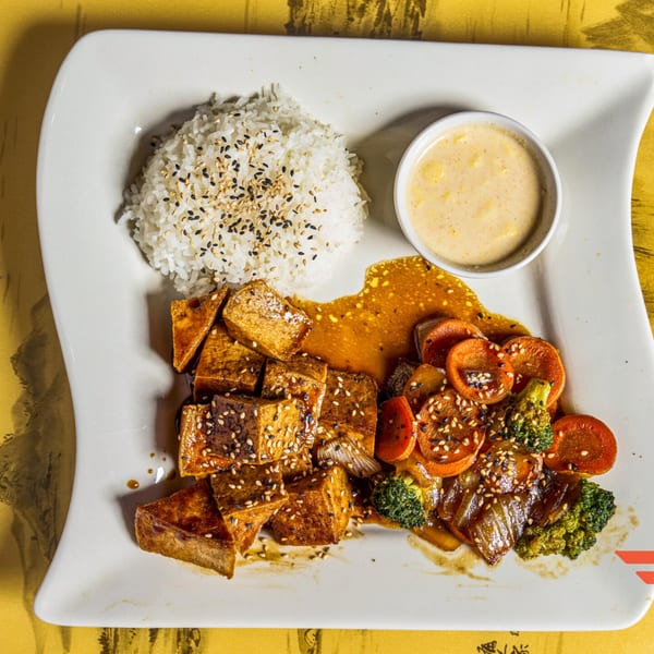 A white plate with sesame-crusted tofu, rice, stir-fried vegetables, and a side of sauce.