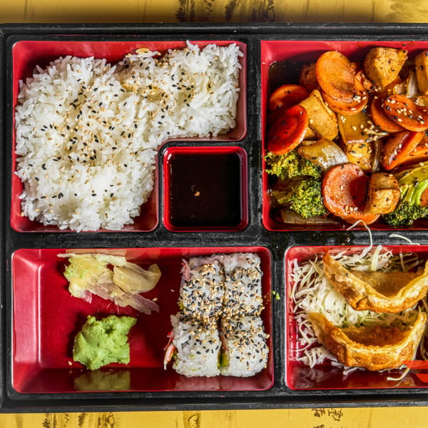 Overhead view of a bento box with rice, stir-fried vegetables, sushi, and dumplings.