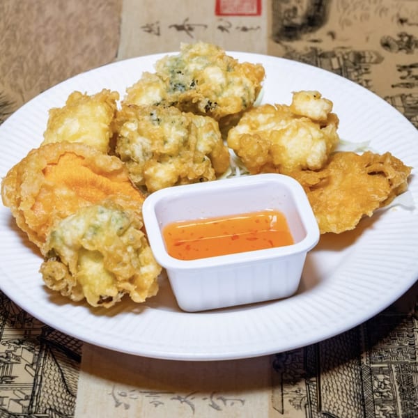 A white plate of golden-fried tempura vegetables and seafood with a side of orange dipping sauce.