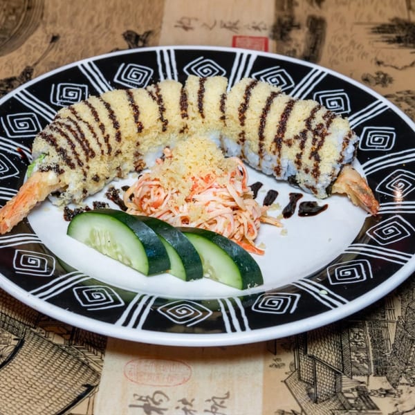 Tempura shrimp sushi roll with eel sauce, crab salad, and cucumber slices on a black and white plate.