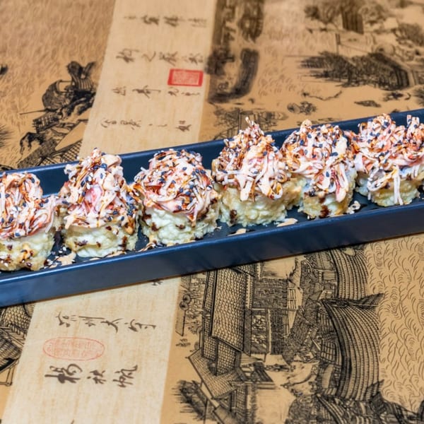 A black tray holds six pieces of sushi topped with sauce and sesame seeds on a patterned surface.